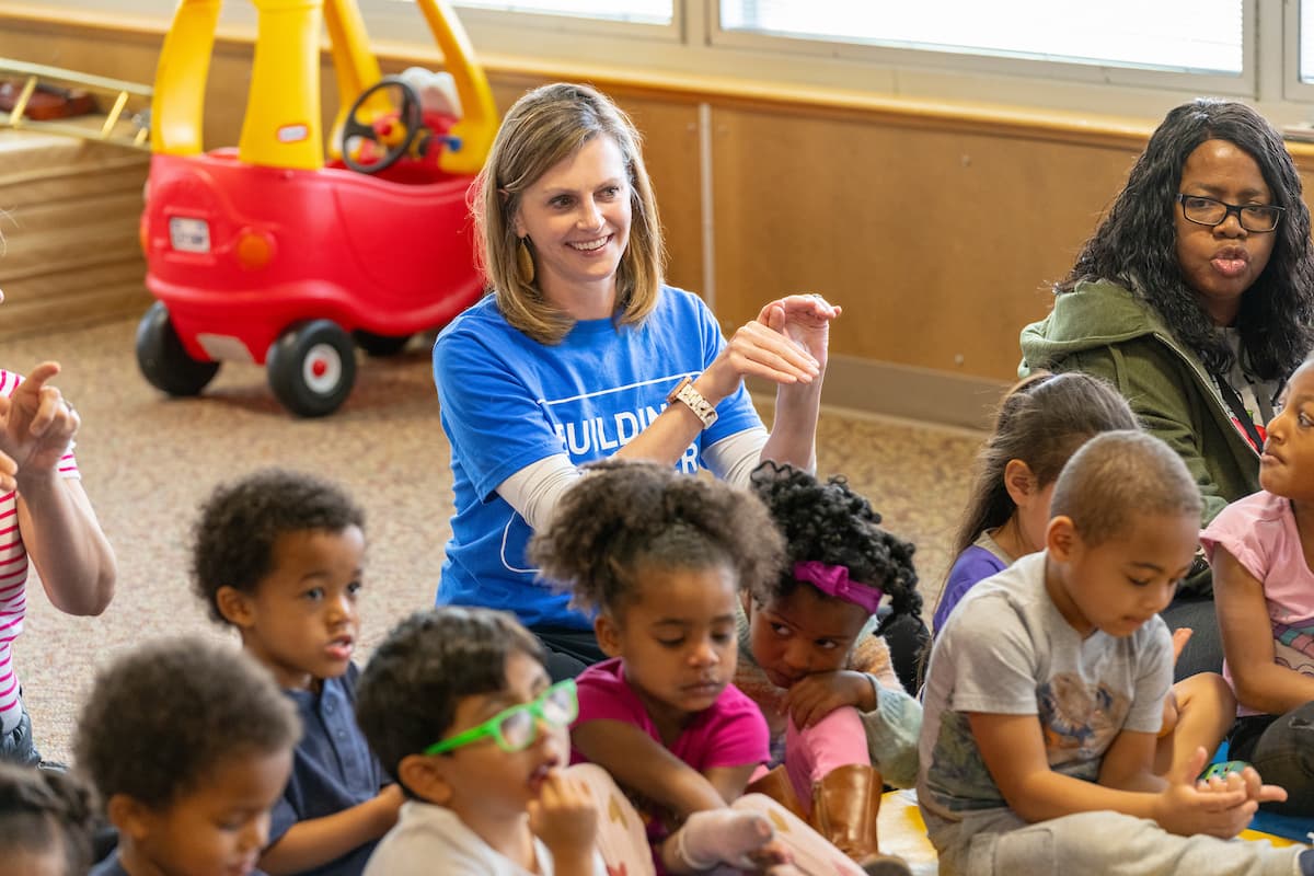 A volunteer in a blue Building Up T-shirt sits on a classroom floor with a group of preschool children