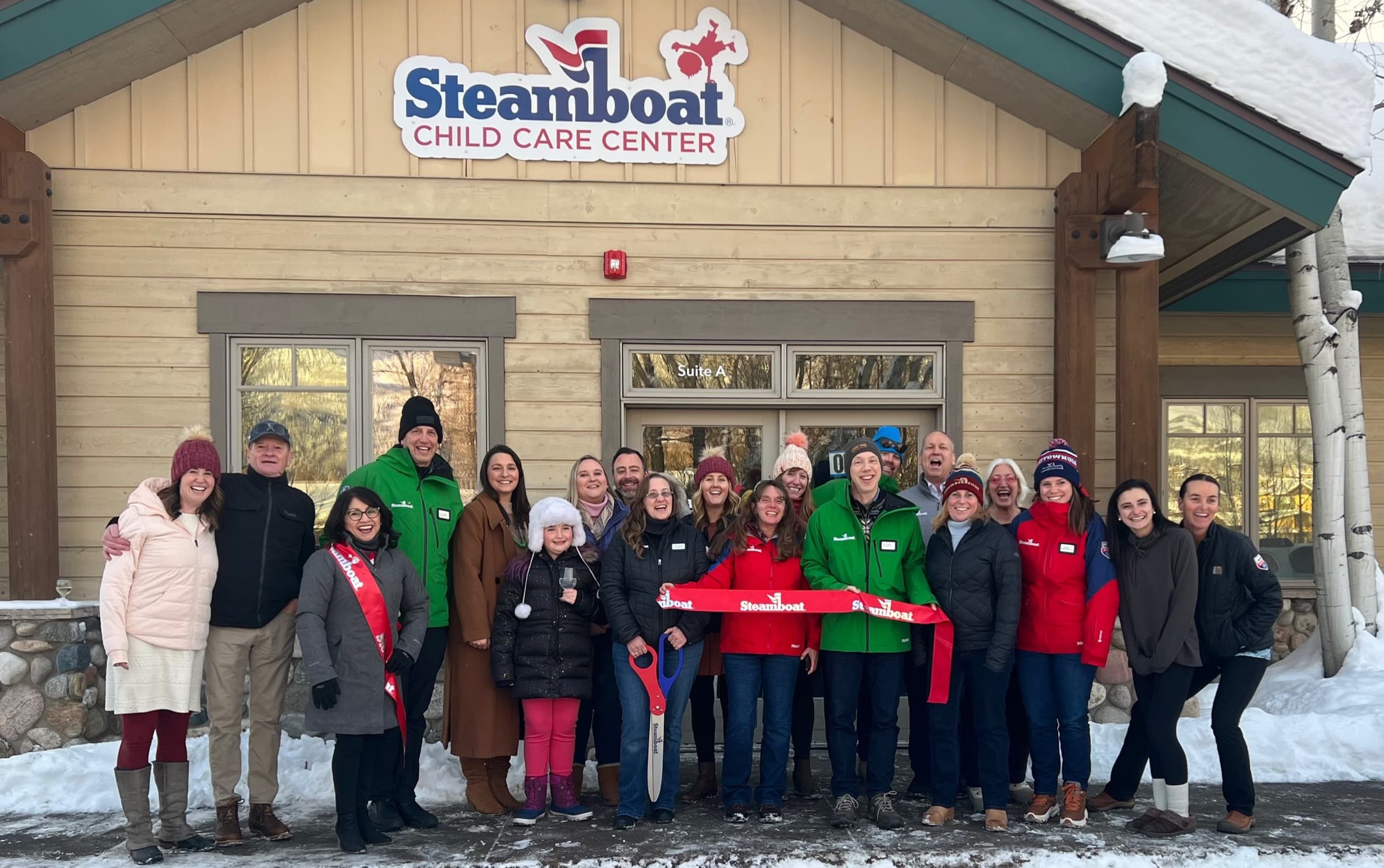 About 25 people in winter coats hold a red ribbon outside the Steamboat Child Care Center in the snow